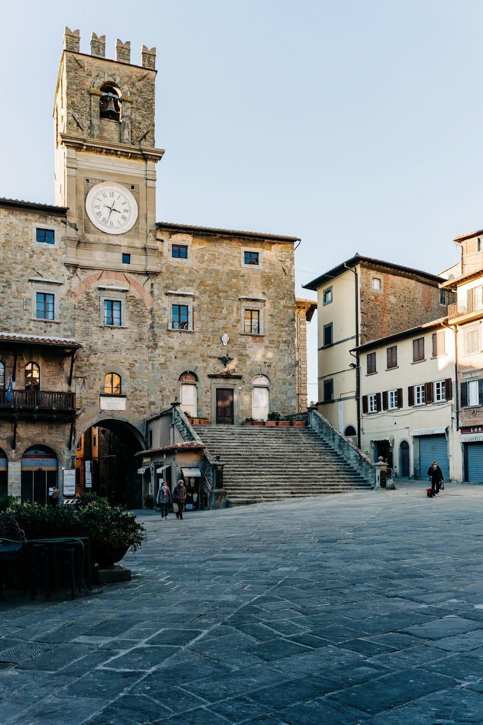 Piazza italiana illuminata dal sole con torre dell'orologio e palazzi in pietra.
