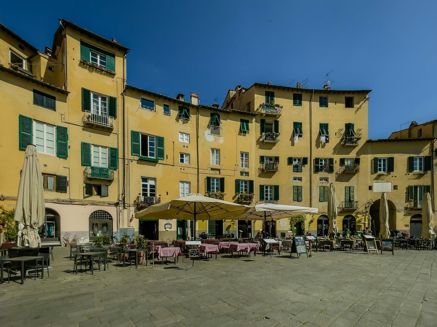 Tavoli vuoti e ombrelloni in una piazza italiana illuminata dal sole.